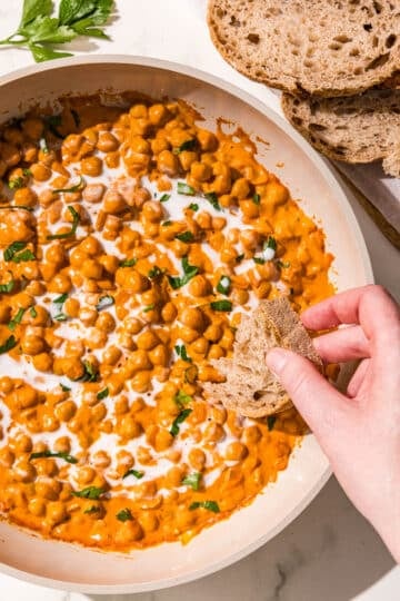 A white pan filled with curry chickpeas topped with fresh cilantro and a drizzle of coconut milk. A white hand is dipping a piece of bread into the chickpeas. Beside the pan are two slices of bread.