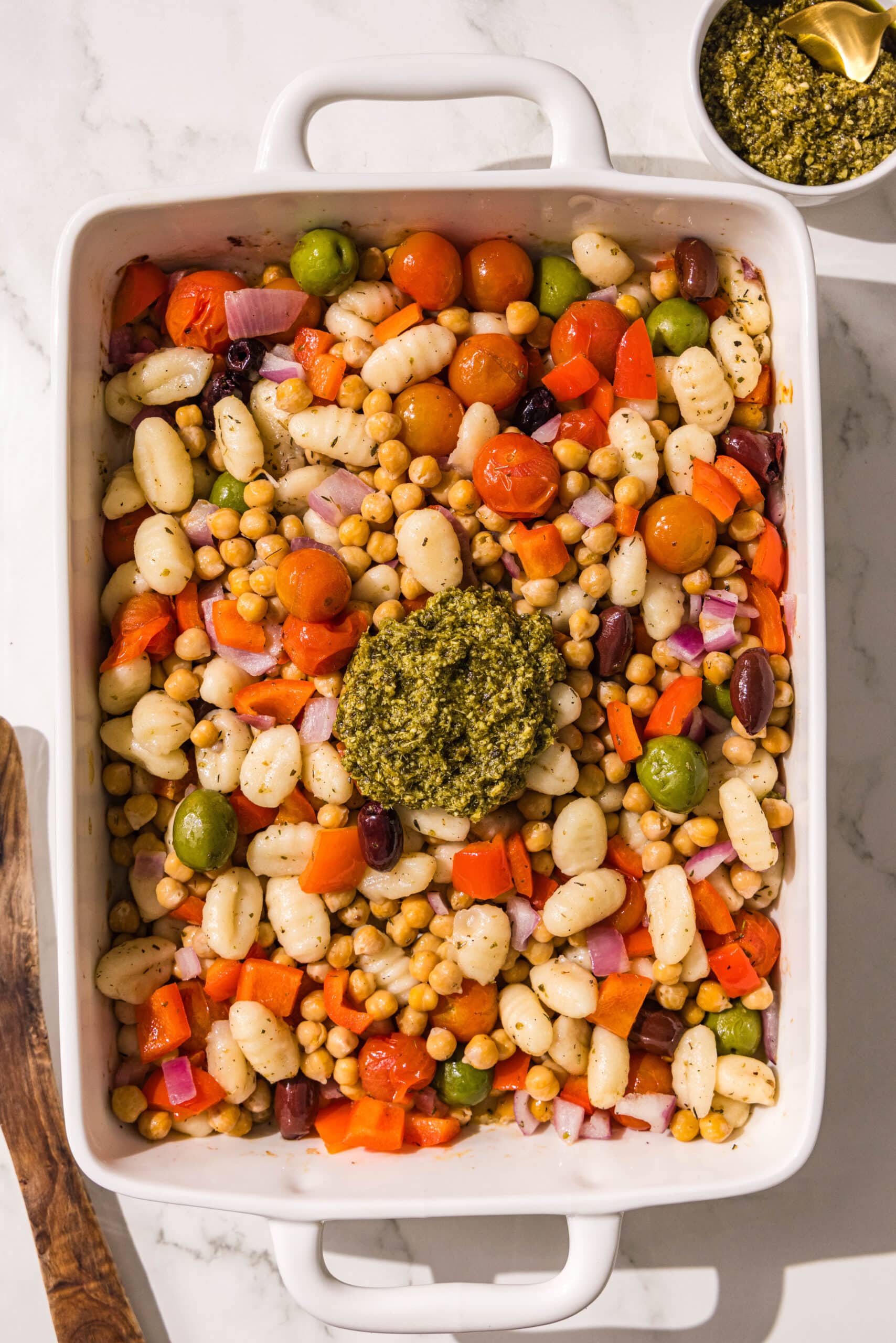 A white casserole dish filled with gnocchi, chickpeas, tomatoes, onions, bell peppers, and olives with a dollop of pesto in the center.