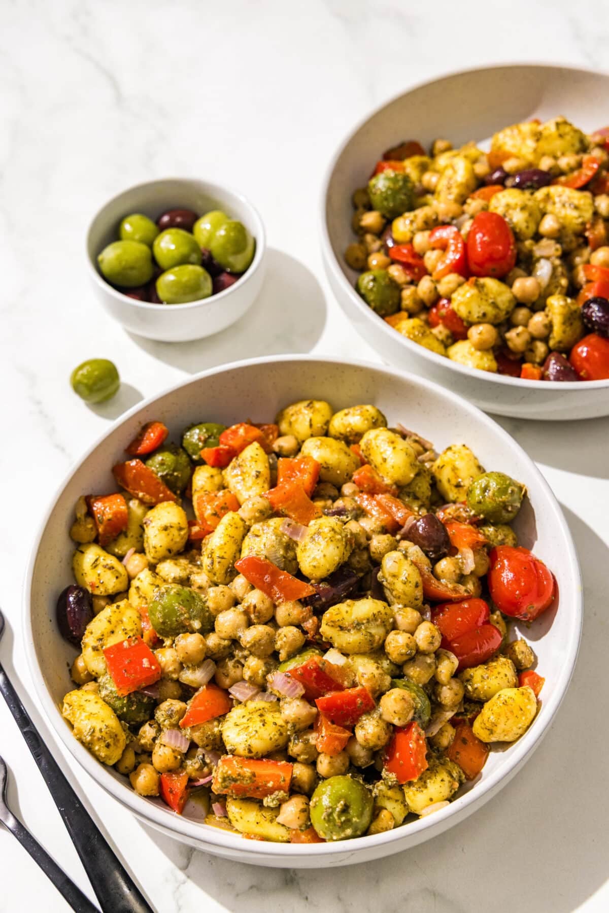 Two bowls of mediterranean pesto gnocchi, a small bowl of olives, and two black metal forks.