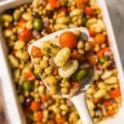 A casserole dish of mediterranean pesto gnocchi and a white spatula holding a scoop of the gnocchi with tomatoes, chickpeas, olives, and gnocchi in it.