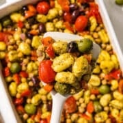 A spatula holding a scoop of pesto mediterranean gnocchi over a casserole dish filled with remaining pesto mediterranean gnocchi.