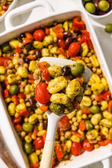 A spatula holding a scoop of pesto mediterranean gnocchi over a casserole dish filled with remaining pesto mediterranean gnocchi.