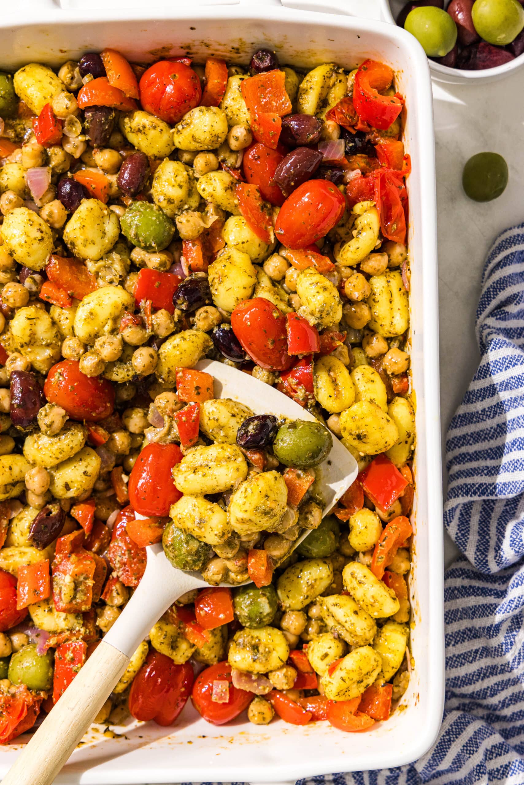 A white casserole dish filled with gnocchi, chickpeas, tomatoes, onions, bell peppers, pesto, and olives with a white spatula in it. Beside the casserole dish is a blue and white striped towel.