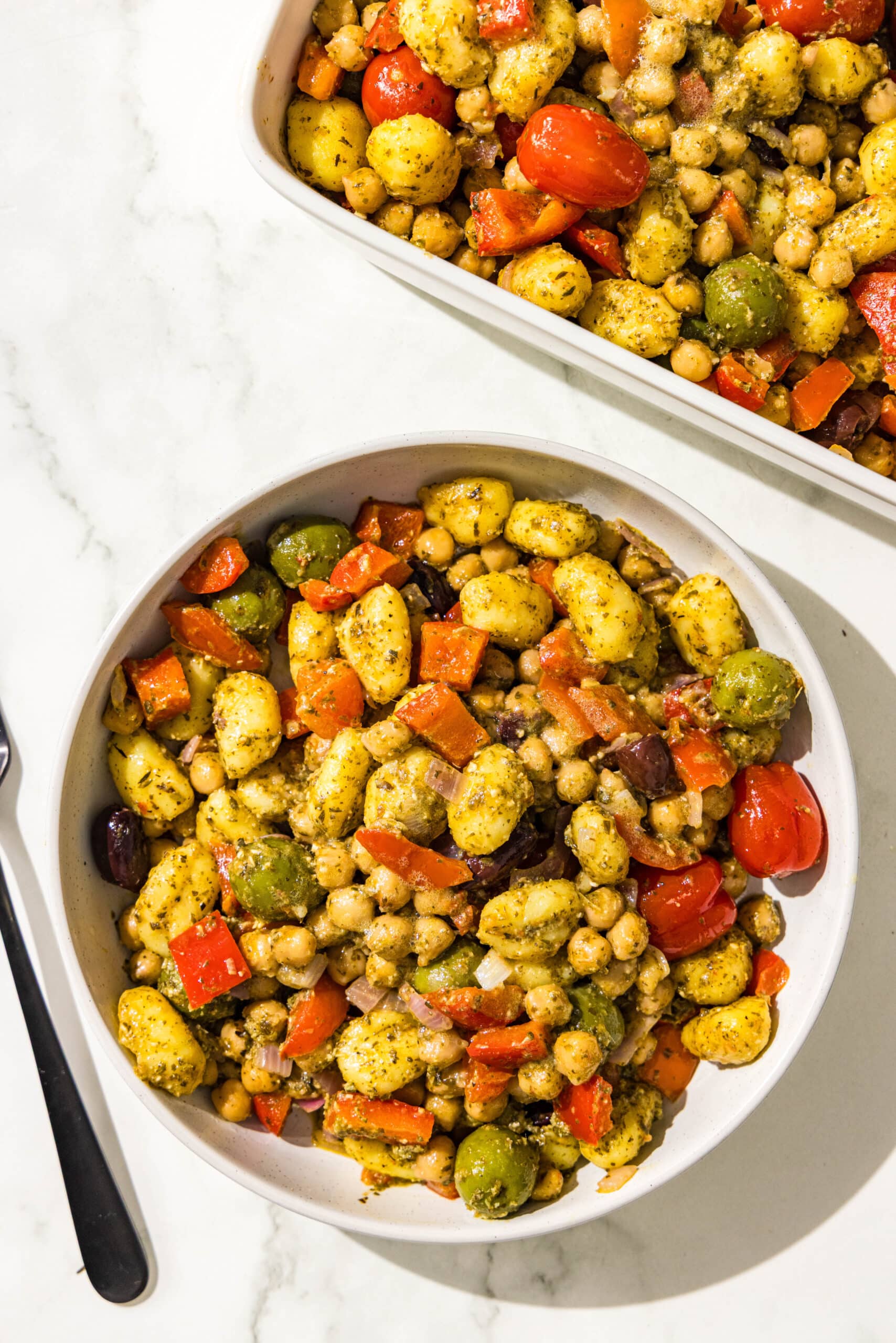 A bowl of mediterranean pesto gnocchi, a casserole dish filled with remaining pesto gnocchi, and a black fork.