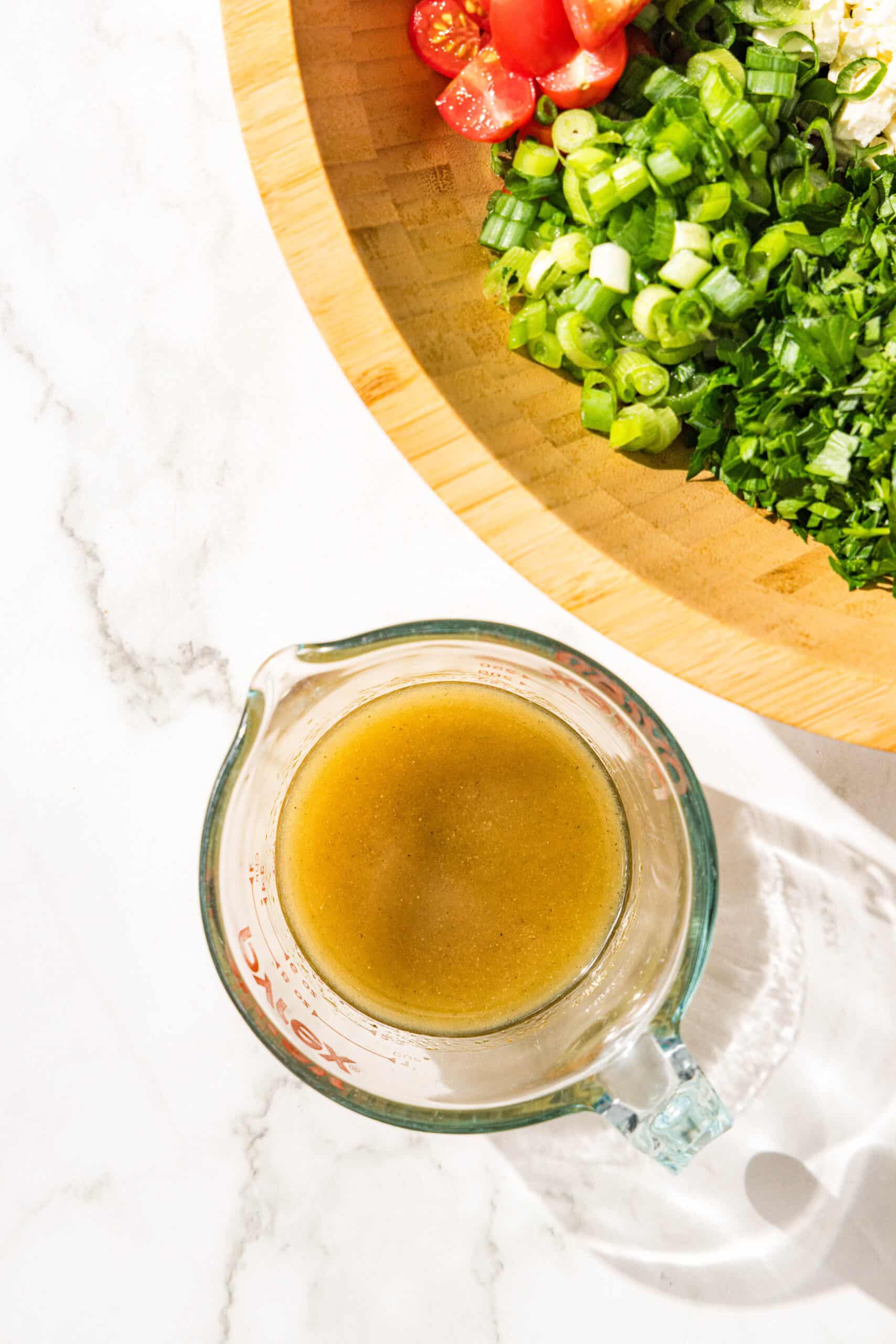 A glass pitcher filled with salad dressing and a wooden salad bowl with bell pepper, tomato, and green onion in it.