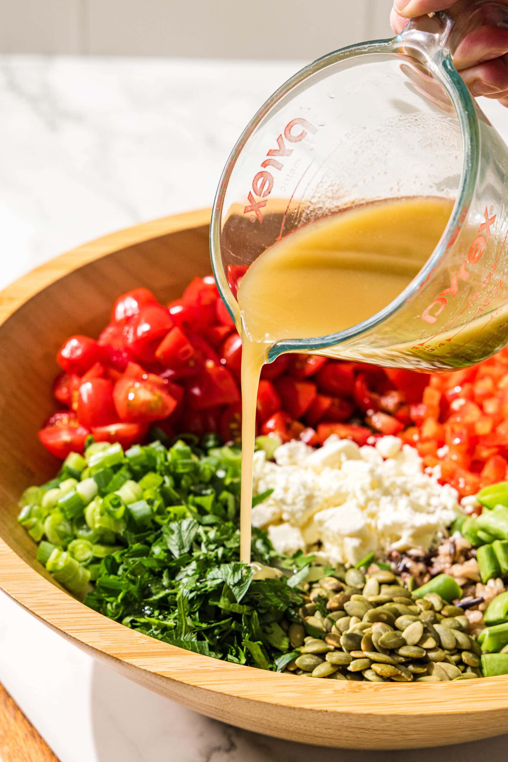 A white hand holding a glass jar pouring salad dressing over a salad in a wooden bowl.