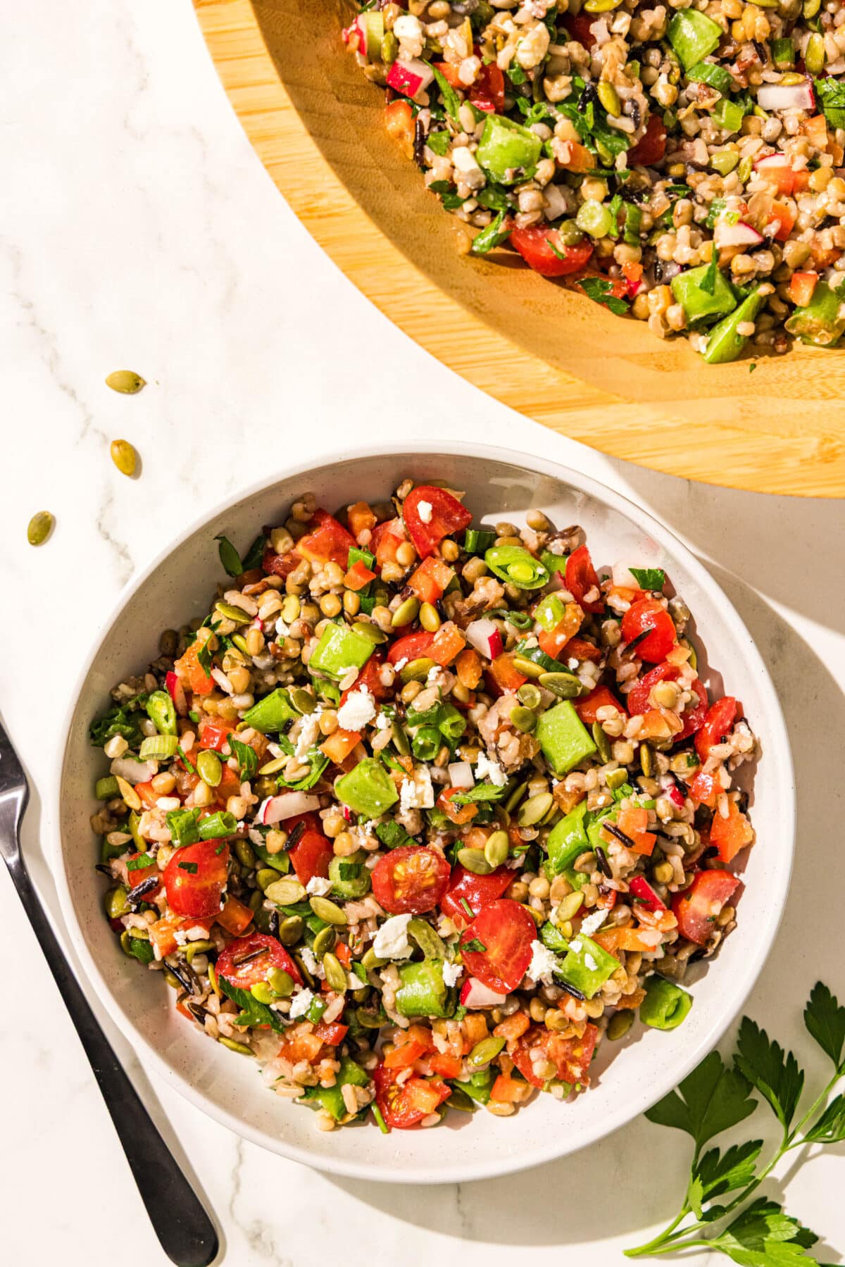 A white glass bowl filled with wild rice lentil salad and a wooden bowl filled with remaining salad. Beside the bowls is a black metal fork and a sprig of fresh parsley.