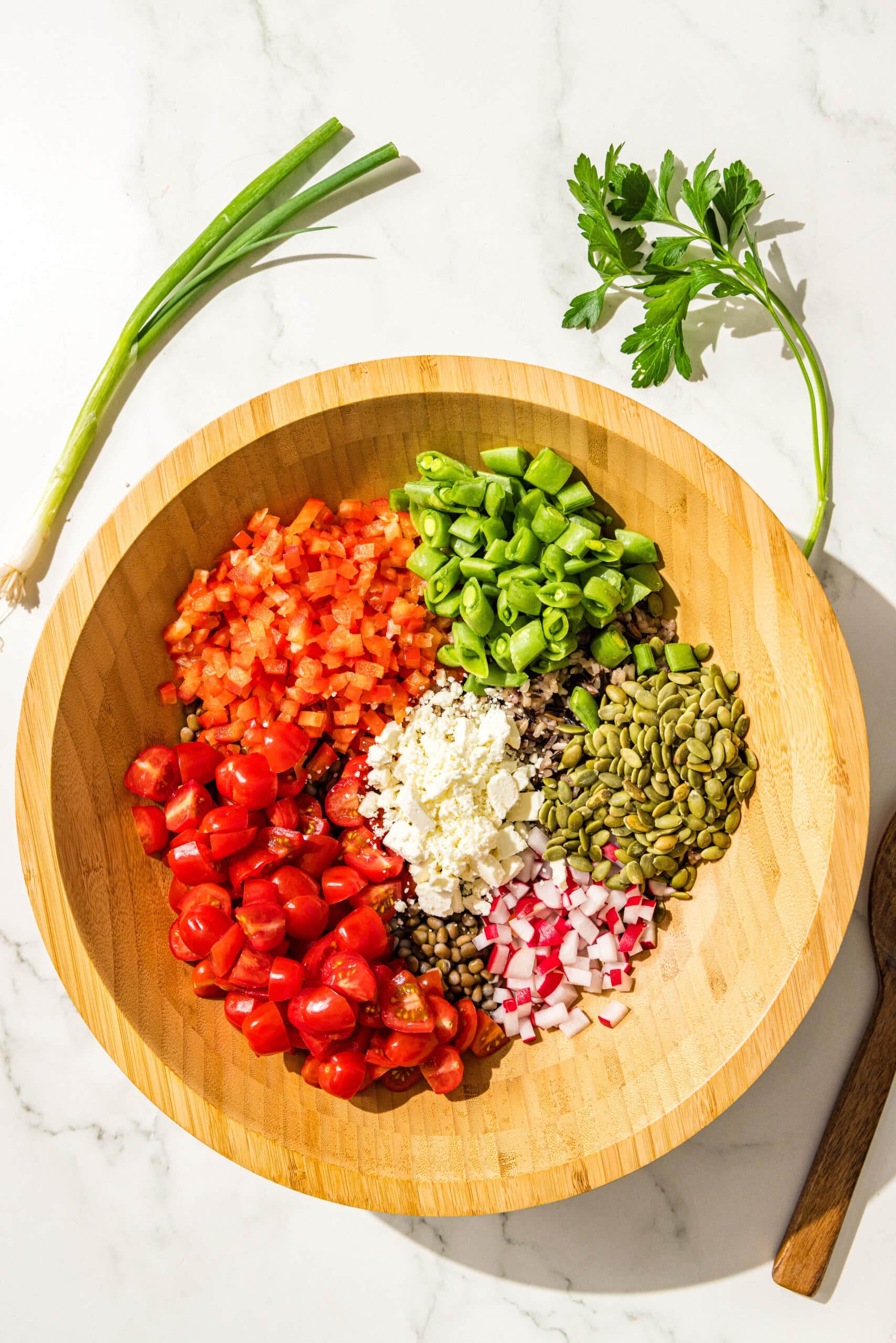 A large wooden salad bowl filled with brown lentils, wild rice, and diced snap peas, tomatoes, bell pepper, radish, pumpkin seeds, and crumbled feta. Beside the bowl is a scallion, a sprig of parsley, and a wooden spoon.