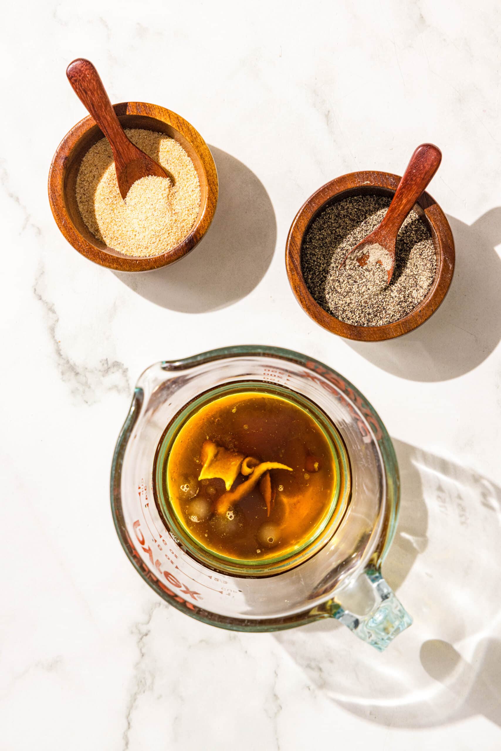 A glass pitcher filled with oil, vinegar, and dijon. Beside the glass pitcher is two small wooden bowls of garlic powder and black pepper.
