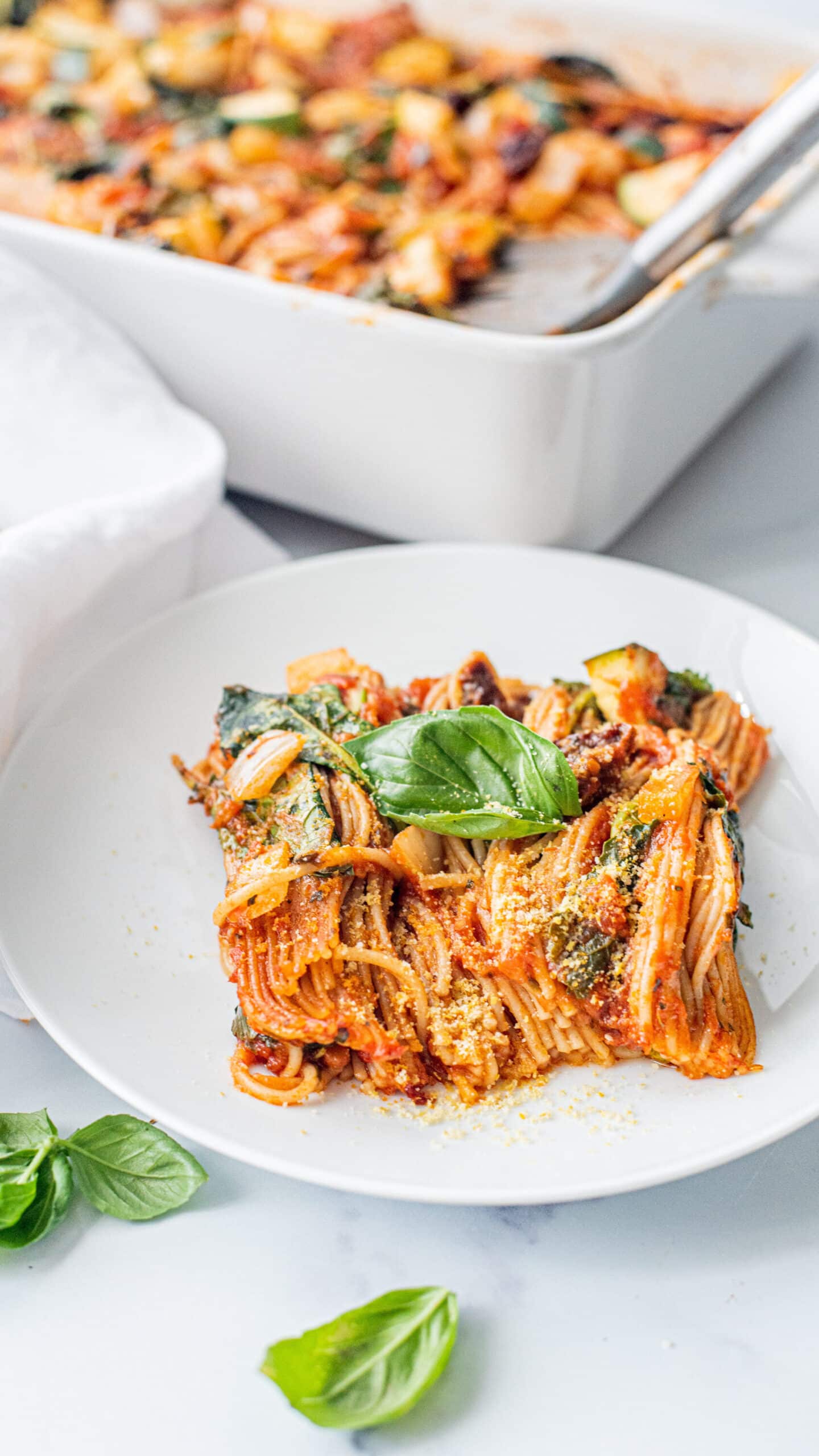 A white plate with a scoop of baked spaghetti on it. In the background is a white 9x13 casserole dish filled with remaining pasta.