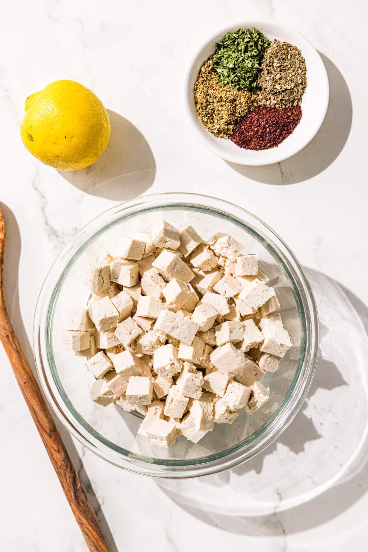 A glass bowl filled with tofu cubes, a wooden spoon, a whole lemon, and a small bowl with spices.