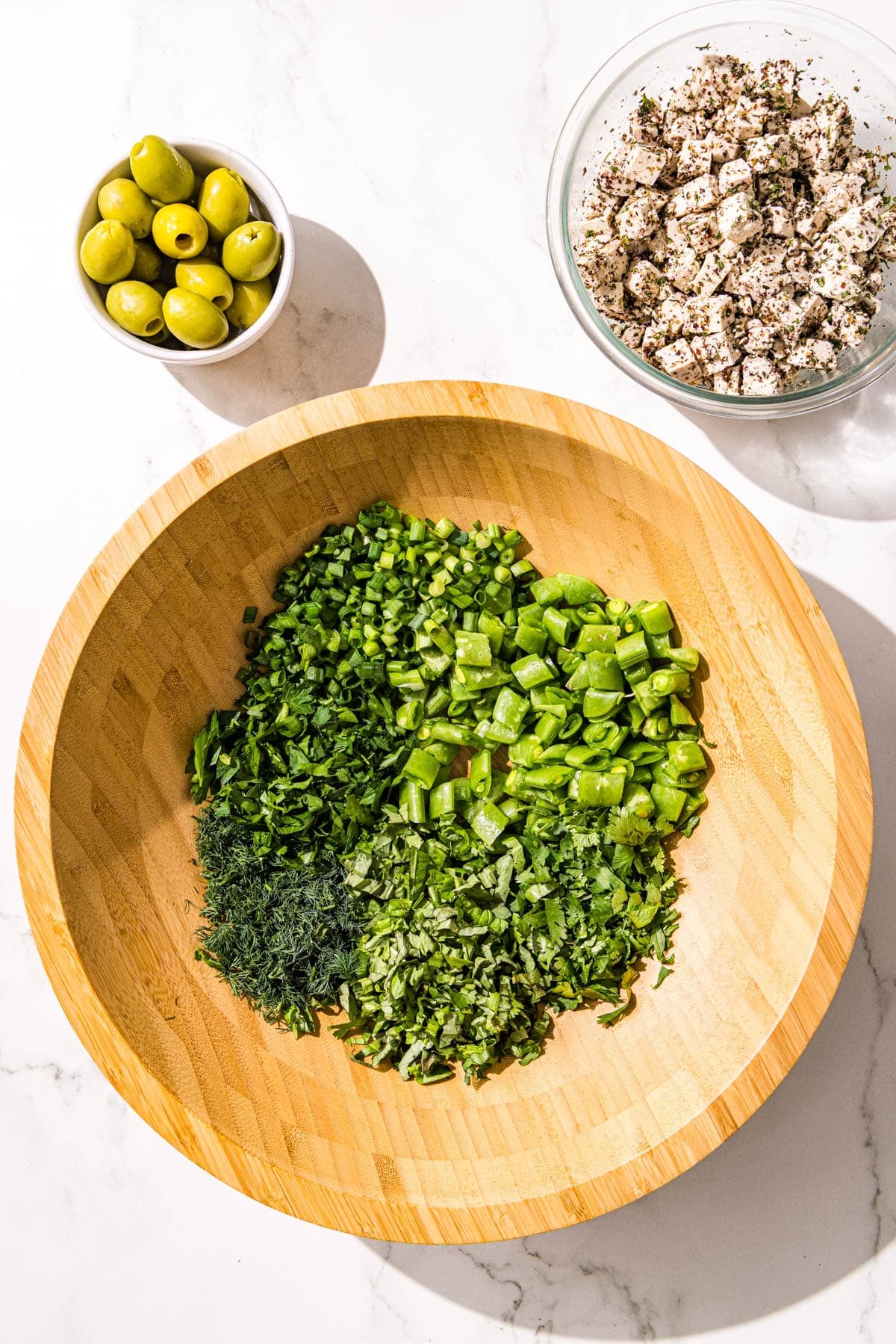 A wooden bowl filled with chopped snap peas, celery, and herbs. Beside the salad bowl is a small of green olives and a glass bowl of tofu.