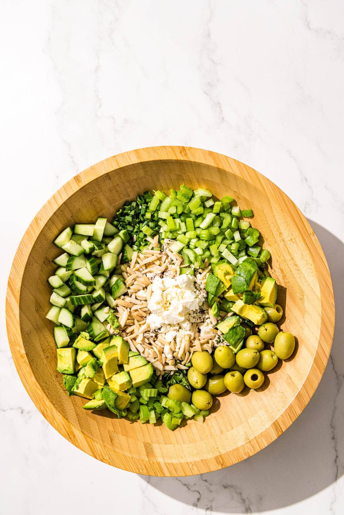 A wooden salad bowl filled with chopped celery, cucumber, green olives, feta, almonds, snap peas, green onion, and fresh herbs.