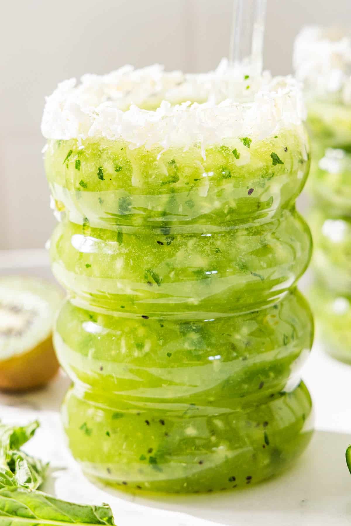 A kiwi lime mocktail in a bubble-shaped glass with a shredded coconut rim and a glass straw sticking out.