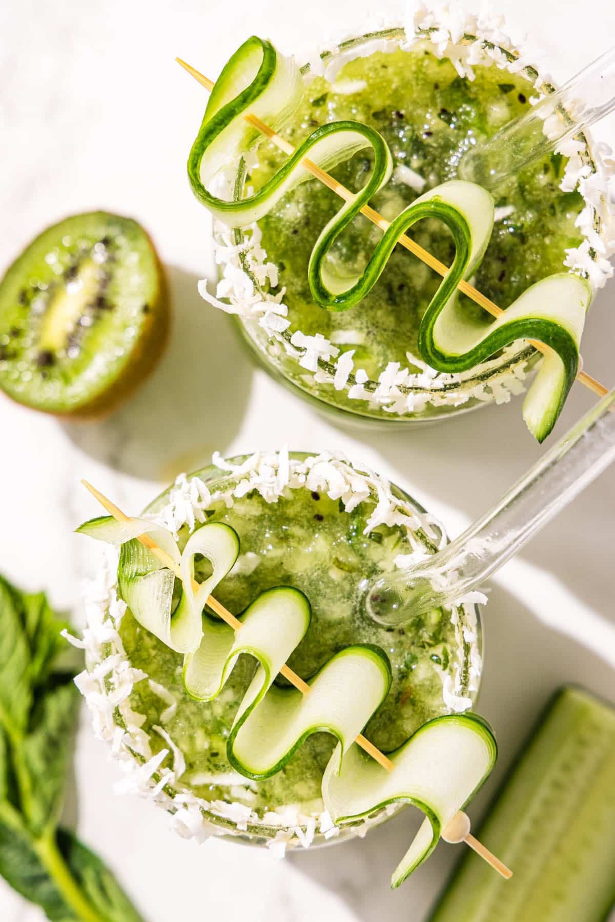 Two kiwi lime mocktails topped with cucumber ribbons on a toothpick in a glass bubble cup. Beside the glasses is half of a kiwi, a slice of cucumber, and a pile of fresh mint.