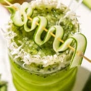 A kiwi lime mocktail in a curved glass topped with a cucumber ribbon on a tooth pick and shredded coconut on the rim. A glass straw sticks out. Beside the glass is half of a kiwi.