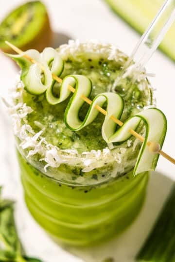 A kiwi lime mocktail in a curved glass topped with a cucumber ribbon on a tooth pick and shredded coconut on the rim. A glass straw sticks out. Beside the glass is half of a kiwi.