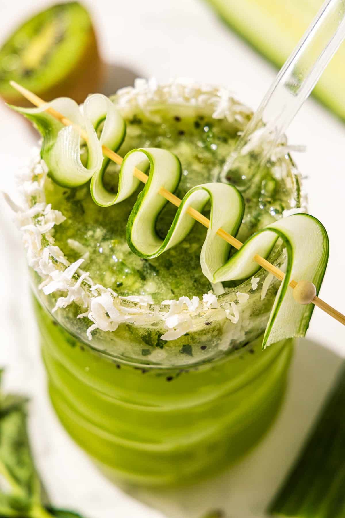 A kiwi lime mocktail in a curved glass topped with a cucumber ribbon on a tooth pick and shredded coconut on the rim. A glass straw sticks out. Beside the glass is half of a kiwi.