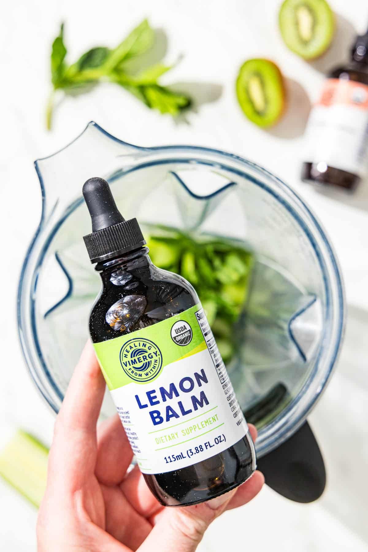 A black dropper bottle with a label on the front that reads "Lemon Balm" and a blender filled with cucumber, kiwi, lime, and mint.