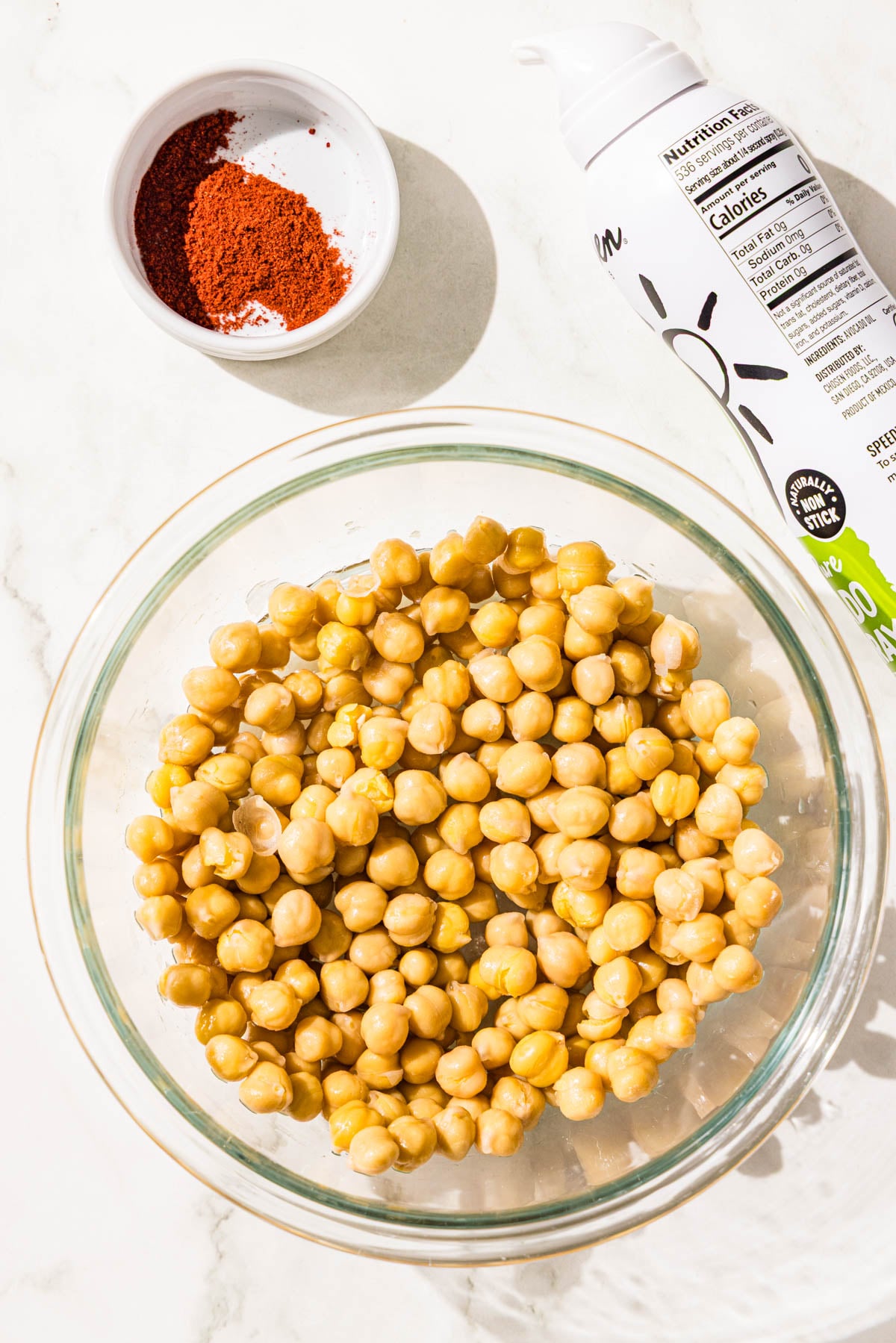 A small glass bowl filled with chickpeas, a bottle of avocado oil spray, and a mini dish of paprika.