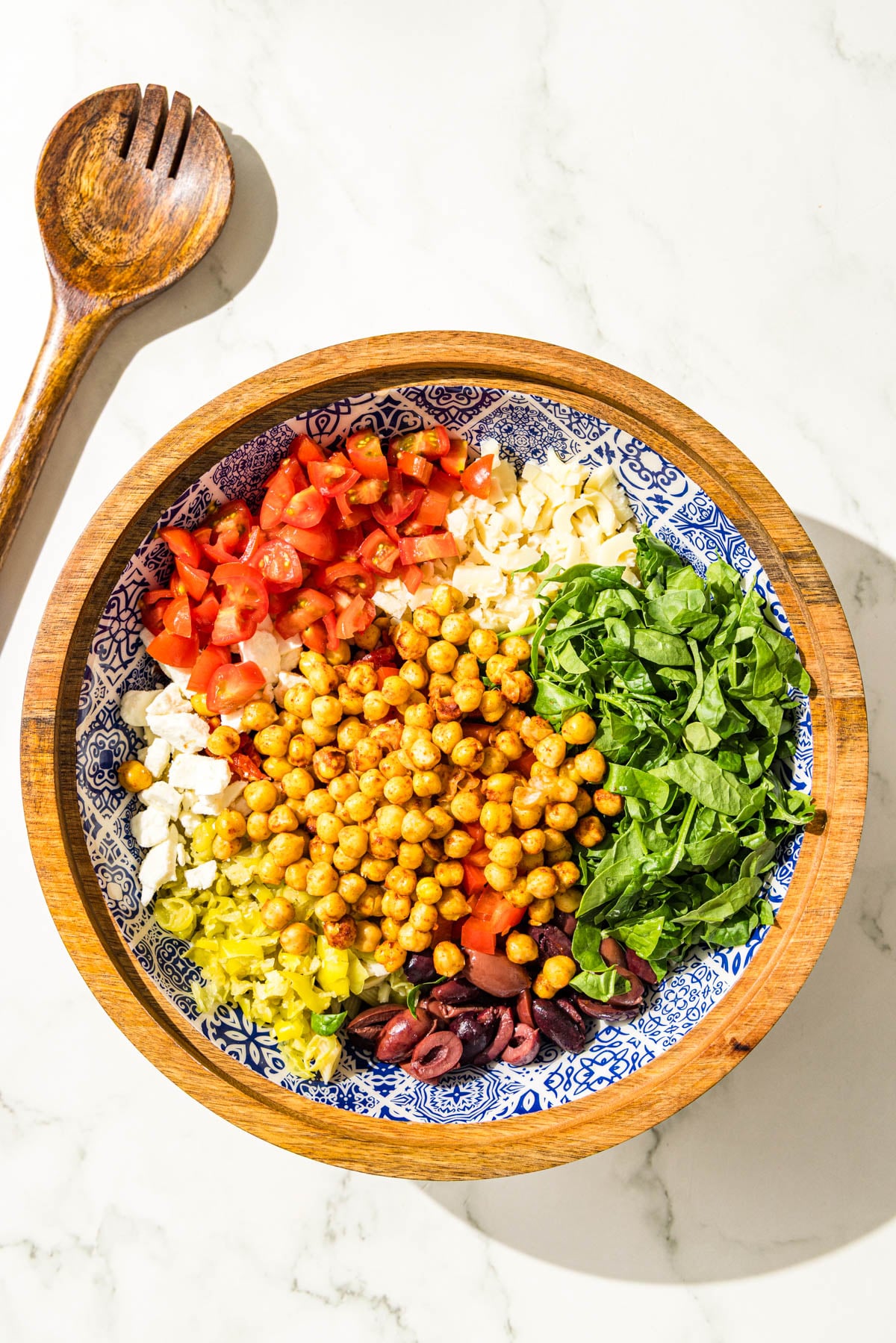 A wooden bowl filled with groups of penne pasta, chickpeas, tomatoes, kalamata olives, spinach, banana peppers, feta, sundried tomatoes and bell pepper.