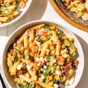Two white glass bowls of pasta salad and a wooden larger bowl with remaining pasta salad plus two black metal forks.