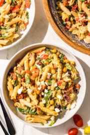 Two white glass bowls of pasta salad and a wooden larger bowl with remaining pasta salad plus two black metal forks.