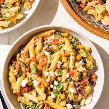 Two white glass bowls of pasta salad and a wooden larger bowl with remaining pasta salad plus two black metal forks.