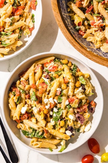 Two white glass bowls of pasta salad and a wooden larger bowl with remaining pasta salad plus two black metal forks.