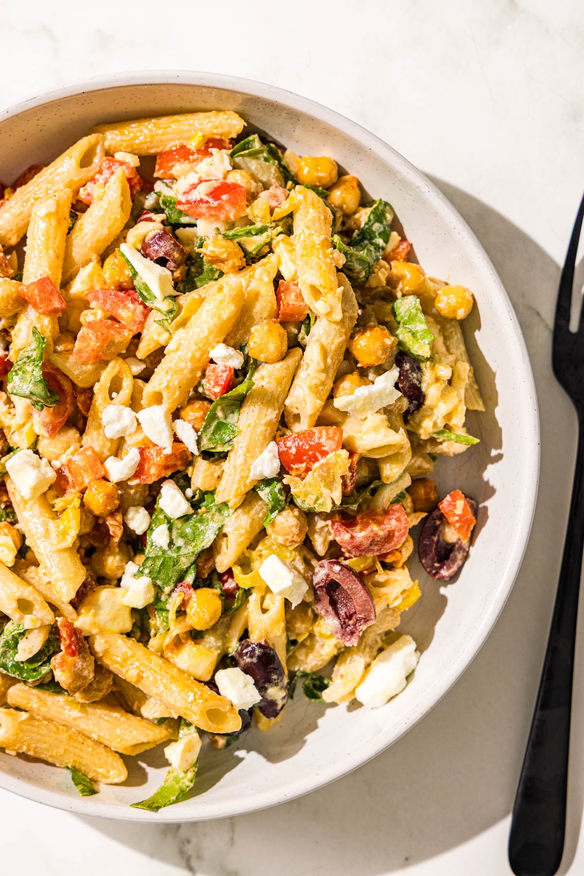 A single white glass bowl of pasta salad topped with feta and a black metal fork beside the bowl.