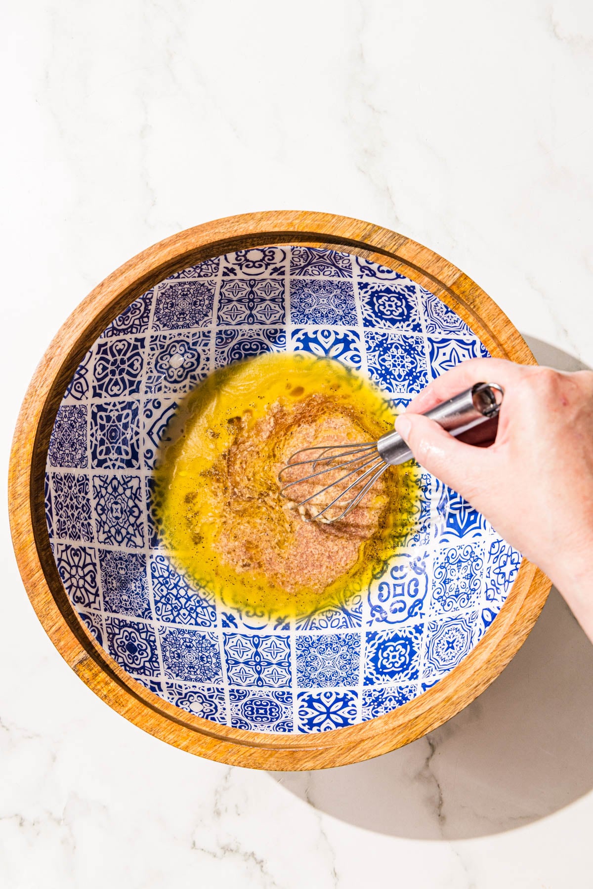 A wooden bowl with tahini and vinegar in the bottom and a white hand holding a mini stainless steel whisk, whisking together the liquids.