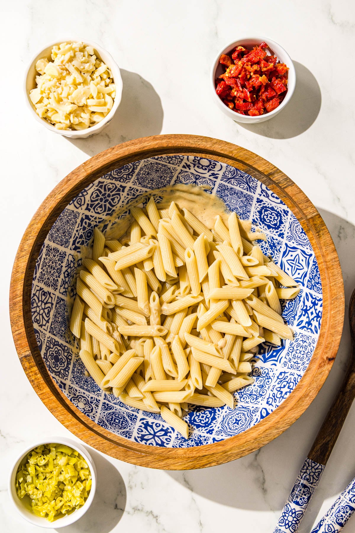 A wooden bowl with tahini dressing and penne noodles, plus three small bowls of hearts of palm, banana peppers, and sundried tomatoes.