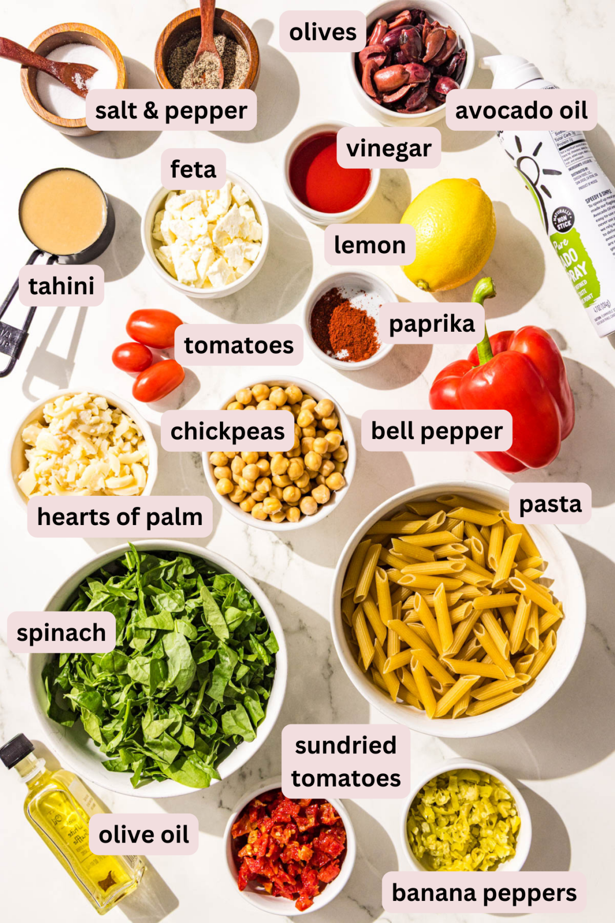 Ingredients laid out on the counter in bowls and labeled including: salt and black pepper, olives, avocado oil spray, feta, vinegar, tahini, lemon, paprika, cherry tomatoes, hearts of palm, pasta noodles, chickpeas, spinach, sundried tomatoes, banana peppers, and olive oil.