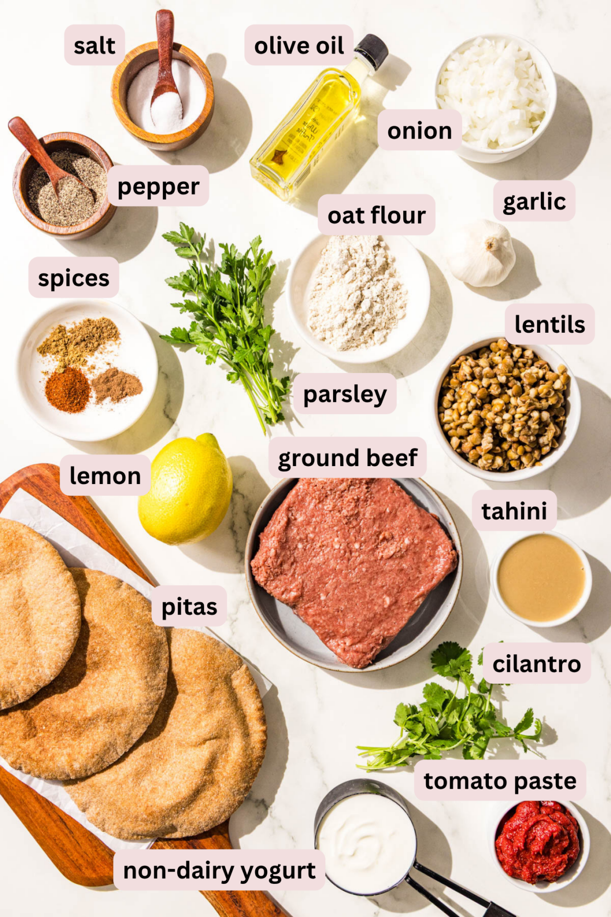 Ingredients laid out on a countertop in bowls and labeled including: salt, black pepper, olive oil, diced onion, garlic gloves, oat flour, fresh parsley, spices, lentils, ground beef, tahini, fresh cilantro, tomato paste, dairy free yogurt, a whole lemon, and pitas.