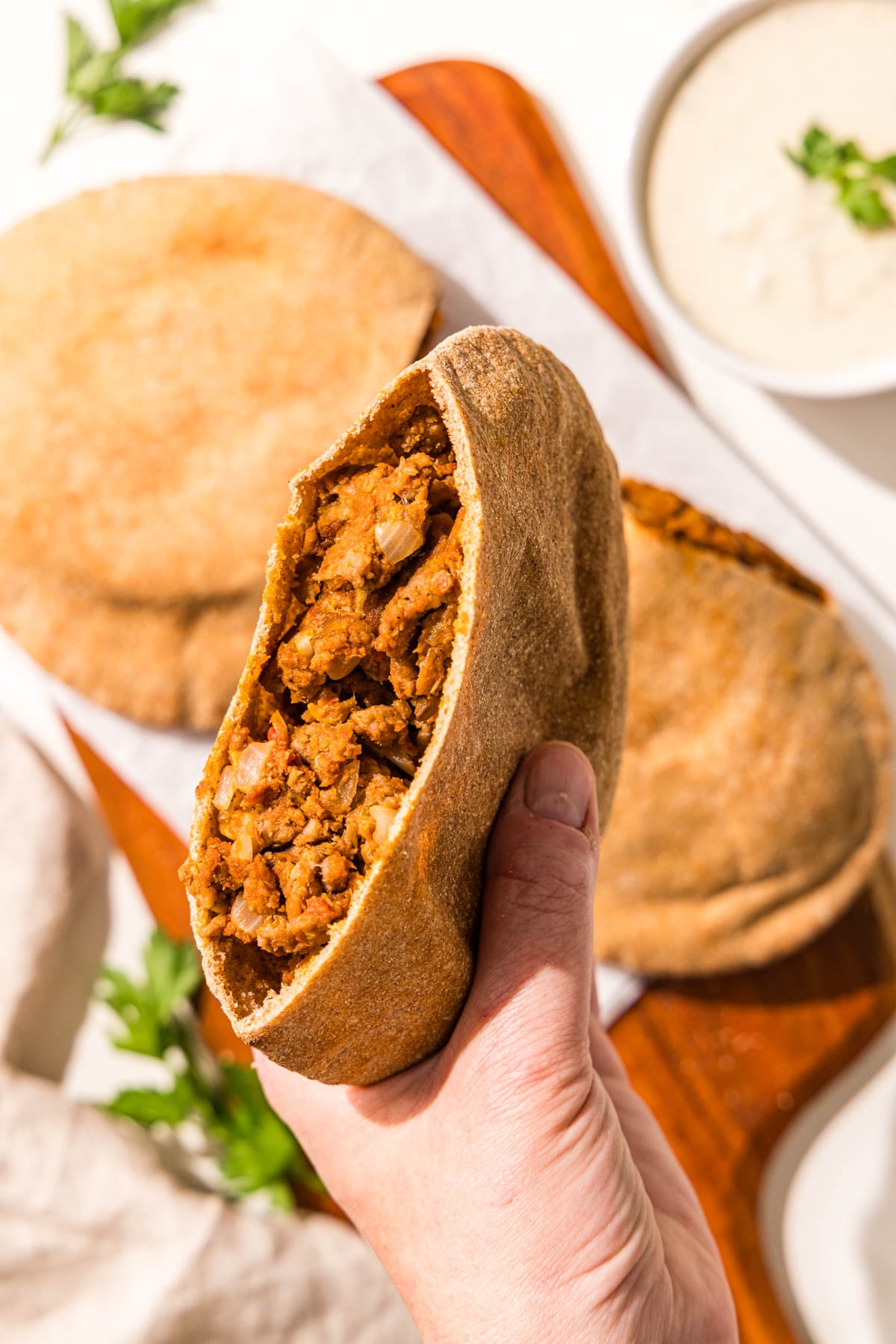 A white hand holding a stuffed pita and a wooden cutting board with two more stuffed pitas on it.