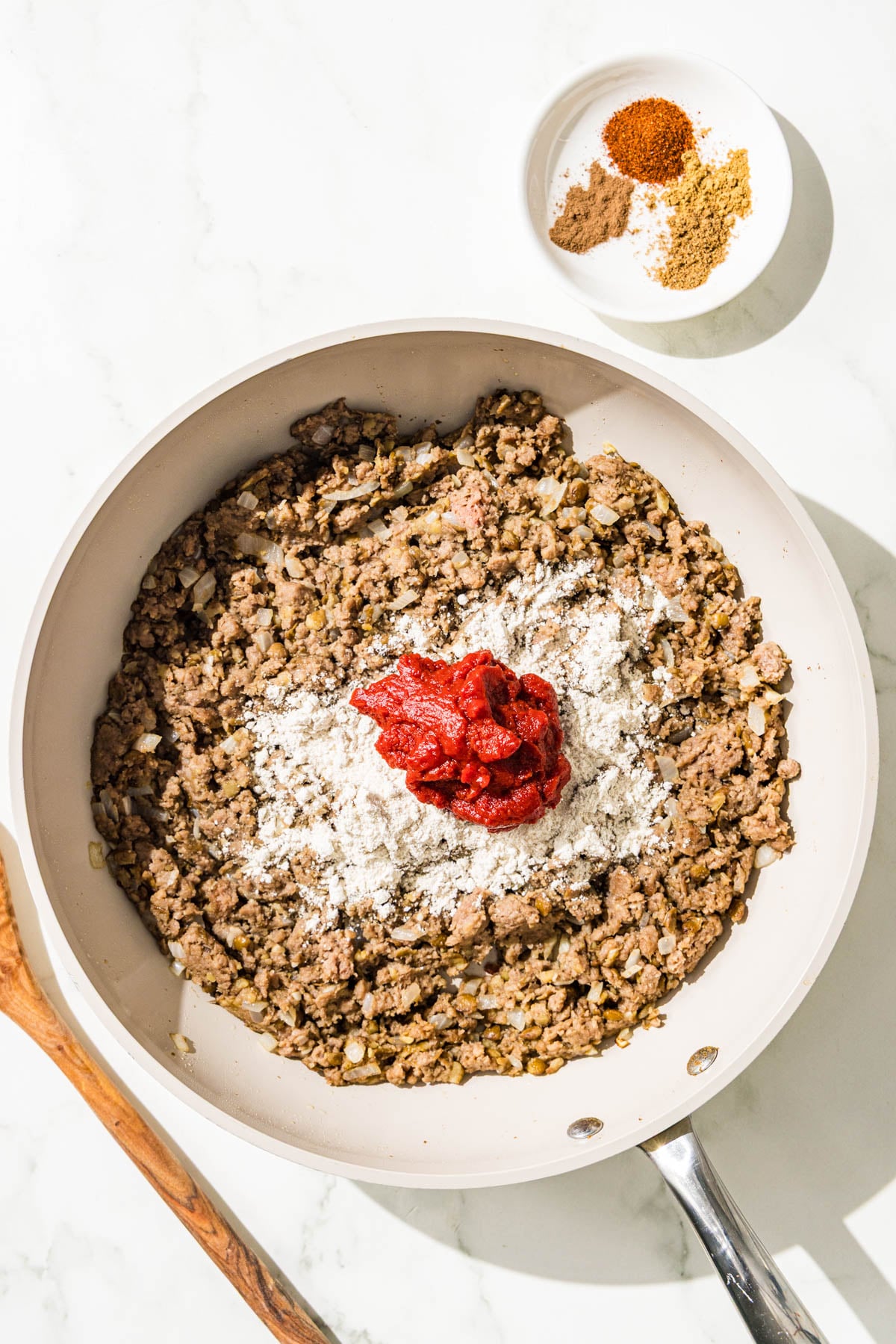 A pan with cooked vegan ground beef and lentils with a scoop of oat flour and tomato paste on top. Beside the pan is a wooden spoon and a small bowl of spices.