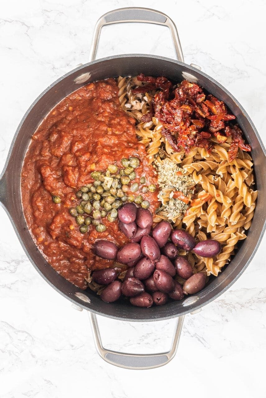 A large pot filled with noodles, marinara, capers, kalamata, olives, sundried tomatoes, and herbs.