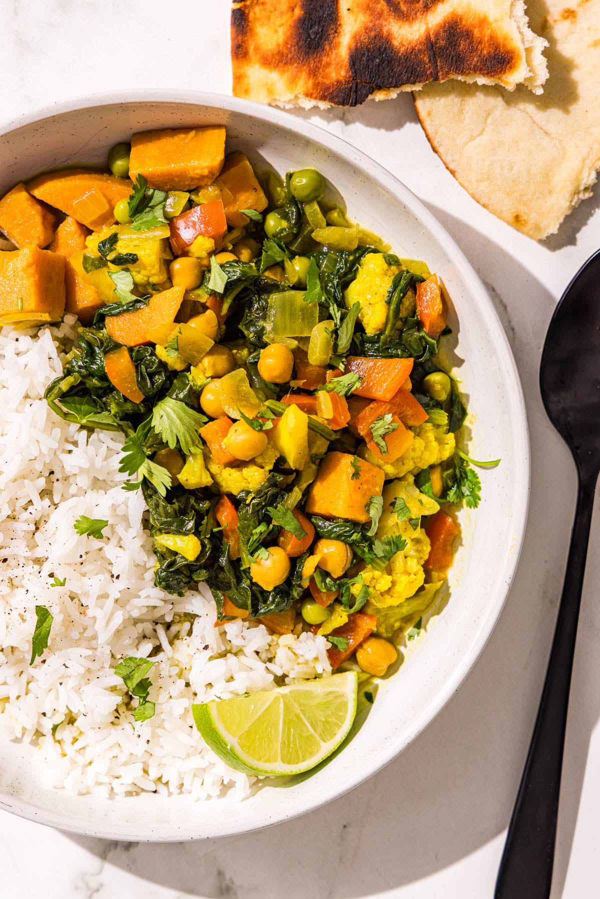 A bowl of sweet potato chickpea curry with white rice, a black metal spoon, and two pieces of naan.