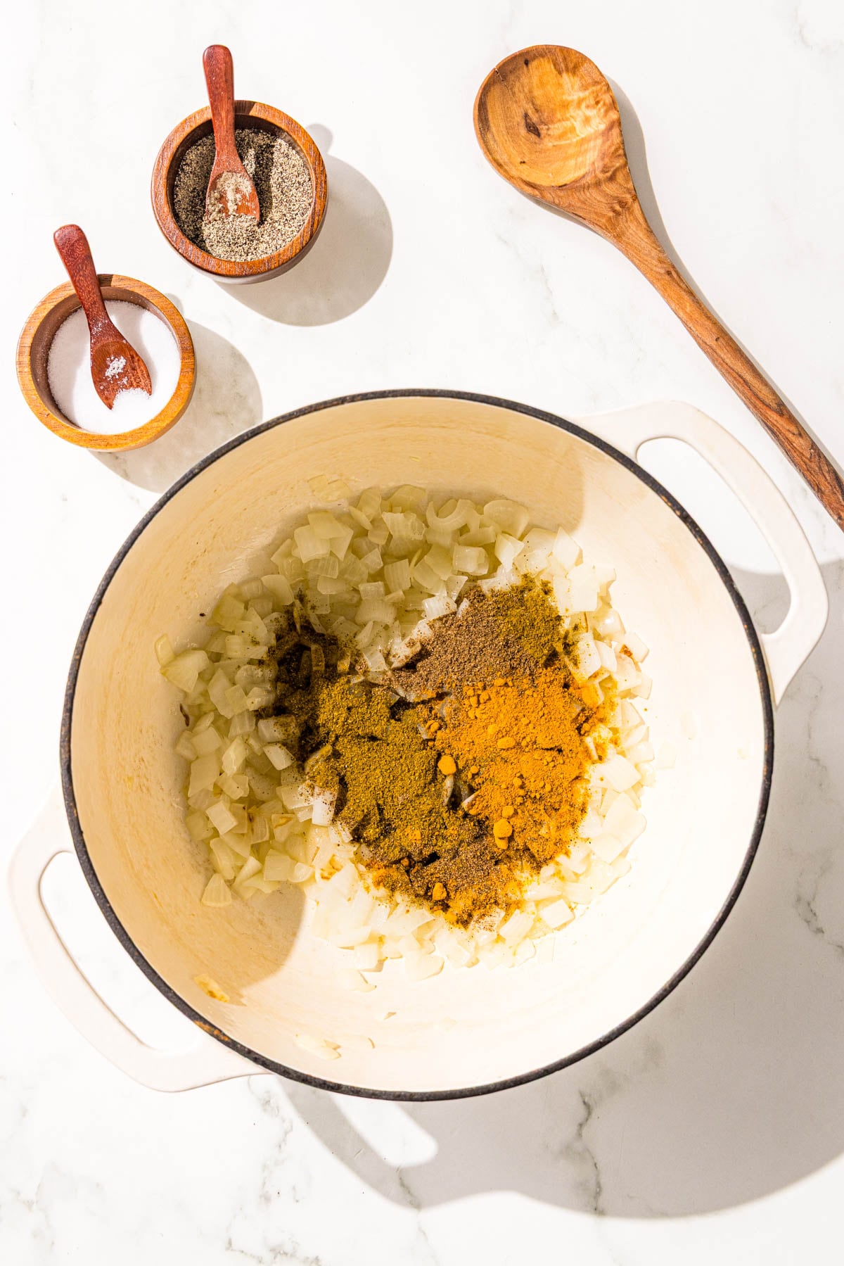A large pot filled with diced onion and spices, along with two small bowls of salt, pepper, and a wooden spoon.