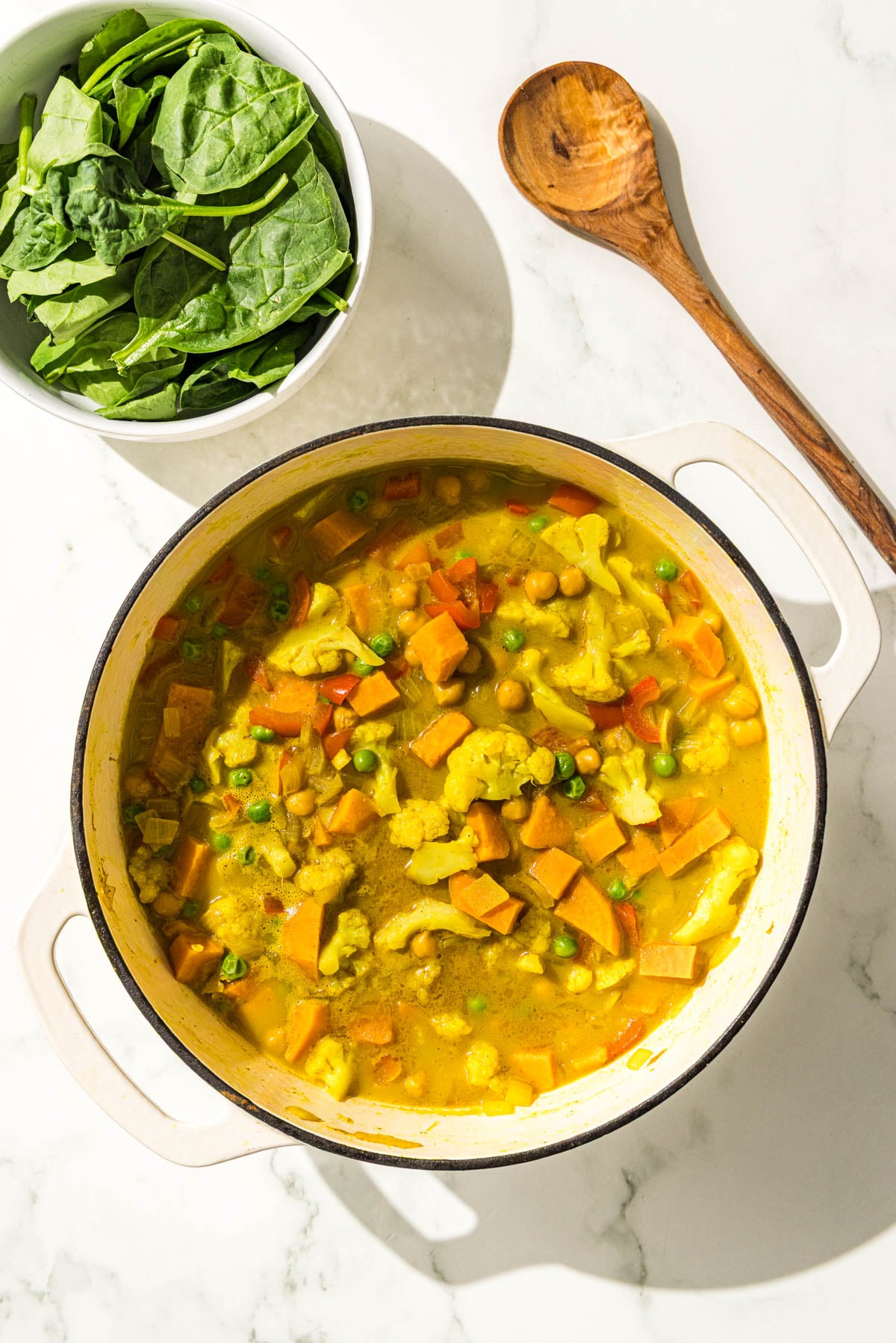 A large pot of sweet potato chickpea curry, a wooden spoon, and a small bowl of fresh spinach.