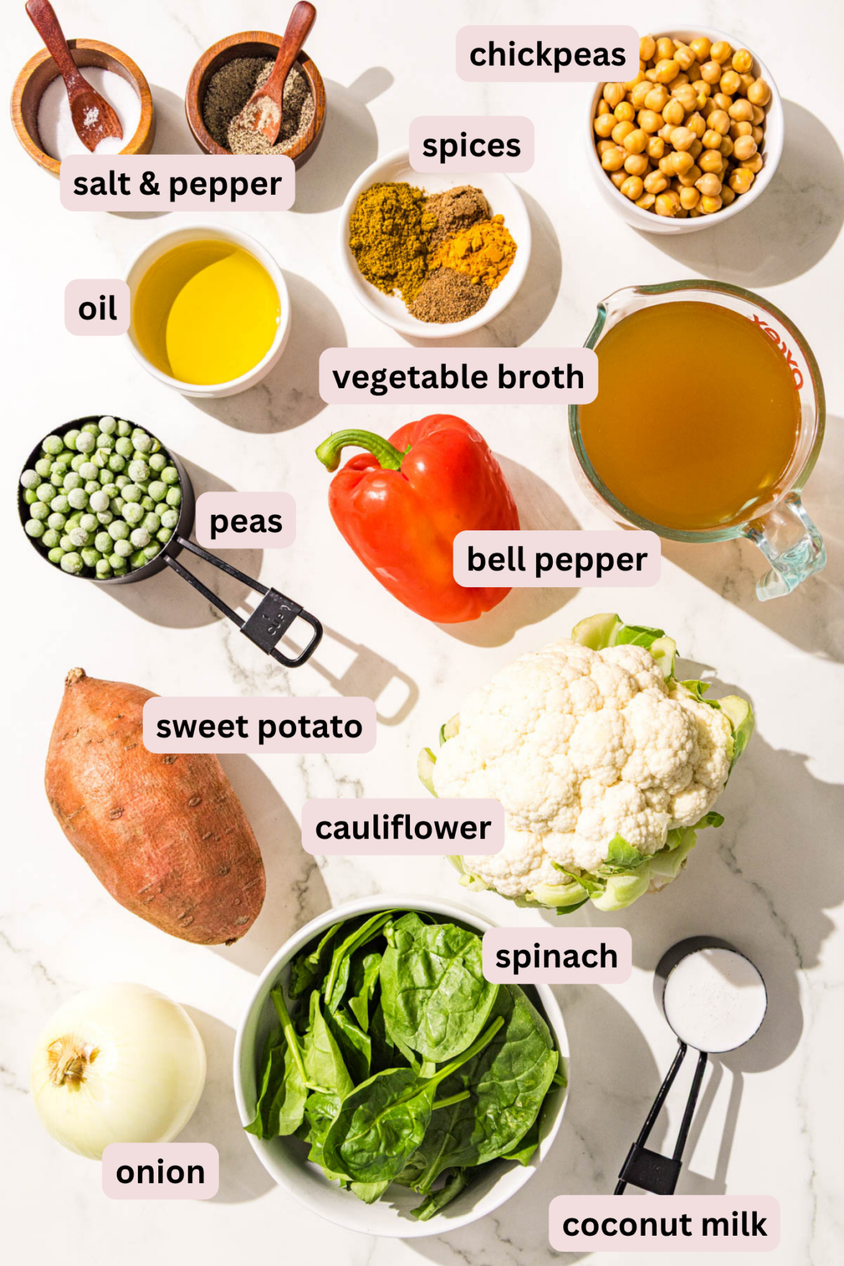 Ingredients needed for sweet potato chickpea curry on the countertop in bowls and labeled including: salt and pepper, chickpeas, vegetable broth, spices, oil, peas, bell pepper, cauliflower, sweet potato, onion, spinach, and coconut milk.