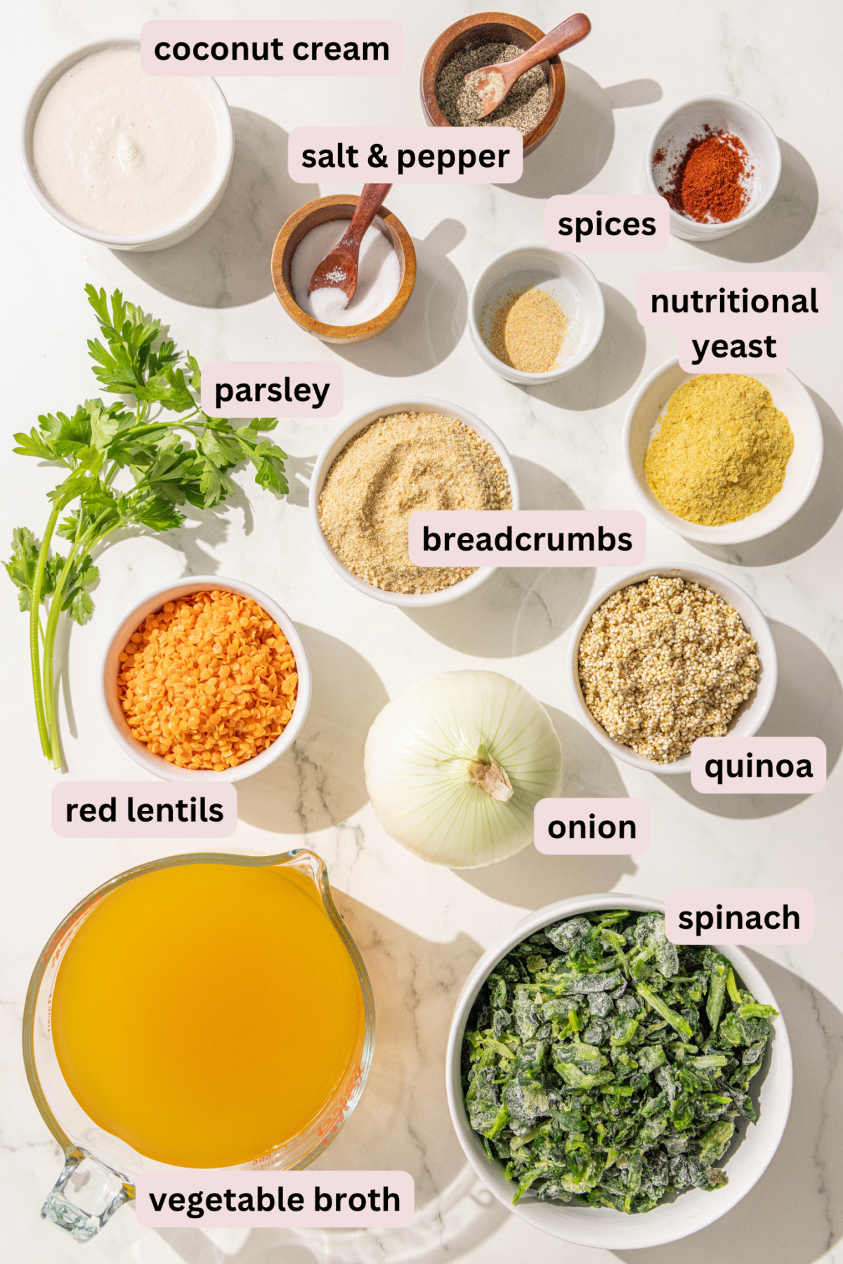 Ingredients laid out on a marble countertop in bowls and labeled including: coconut cream, salt and pepper, spices, nutritional yeast, parsley, breadcrumbs, quinoa, red lentils, onion, spinach, and vegetable broth.