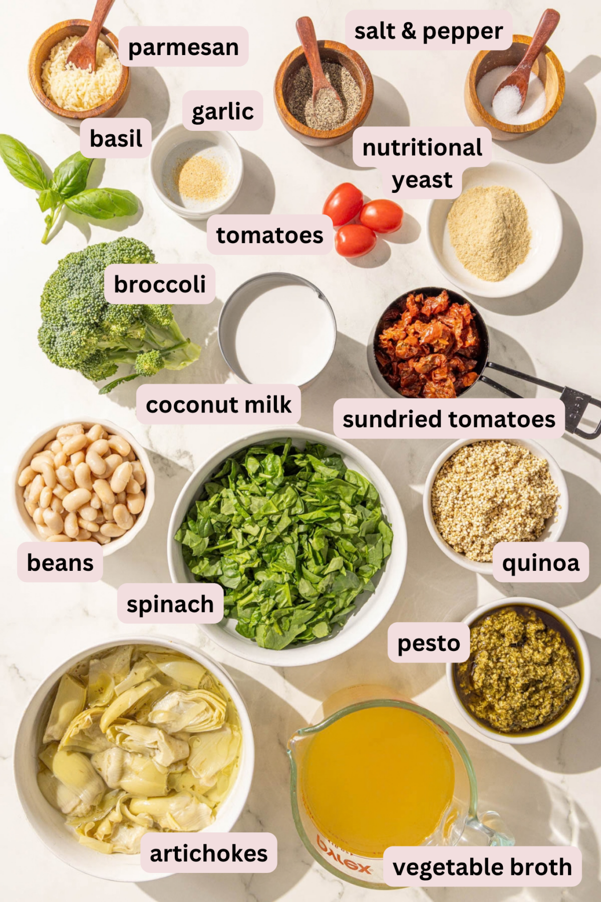 Ingredients laid out on a marble counter top labeled and in bowls including: parmesan, salt and pepper, garlic powder, nutritional yeast, cherry tomatoes, coconut milk, basil, sundried tomatoes, quinoa, white beans, spinach, pesto, artichokes, and vegetable broth.