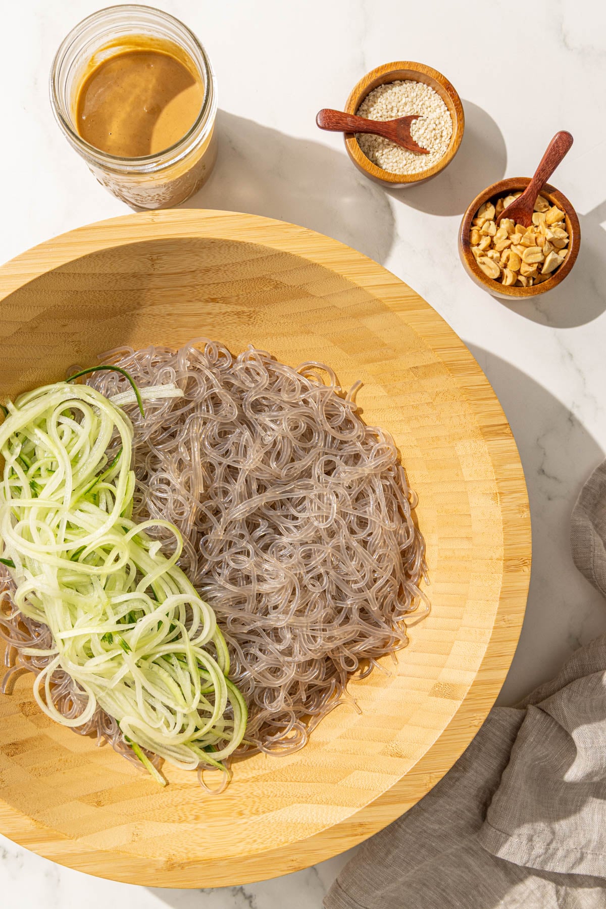 A large wooden bowl of noodles and julienned cucumber and small bowls of peanuts, sesame seeds, and sesame sauce.
