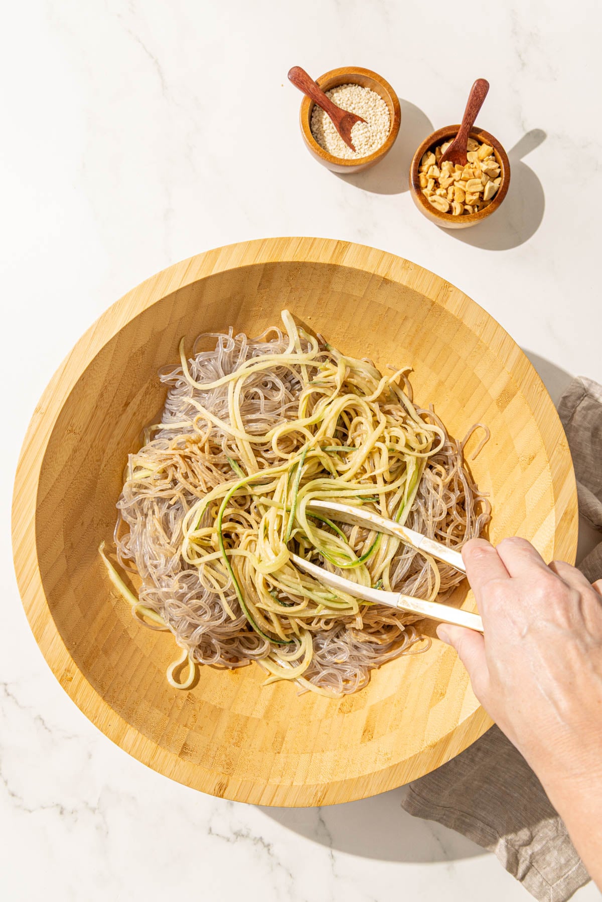 A bowl of noodles, cucumber, and sesame sauce with a white hand holding chopsticks tossing the noodles.
