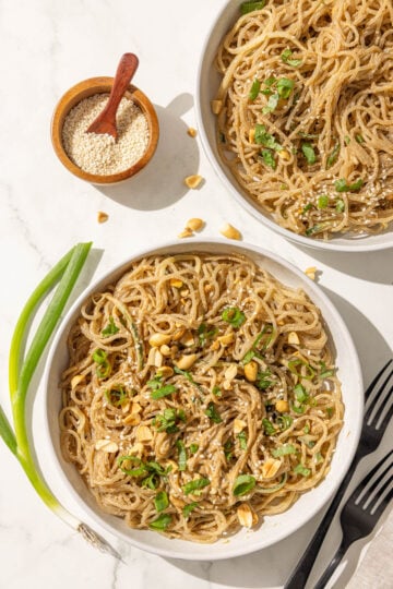 Two bowls of sesame noodles topped with green onion, crushed peanuts, and sesame seeds and then two black forks, a small bowl of sesame seeds, and two green onions.