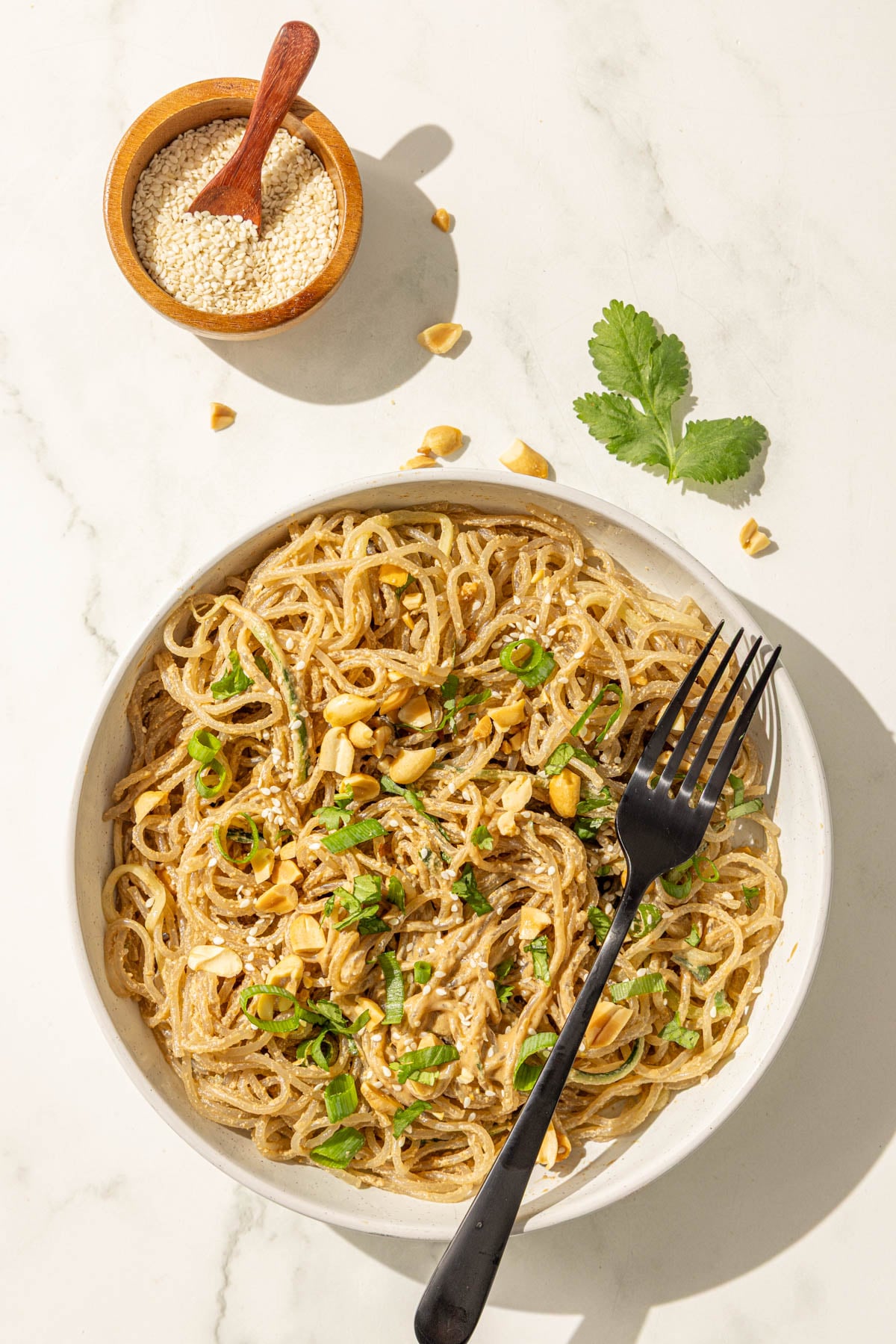 A bowl of sesame noodles topped with peanuts, sesame seeds, green onion and a black metal fork.