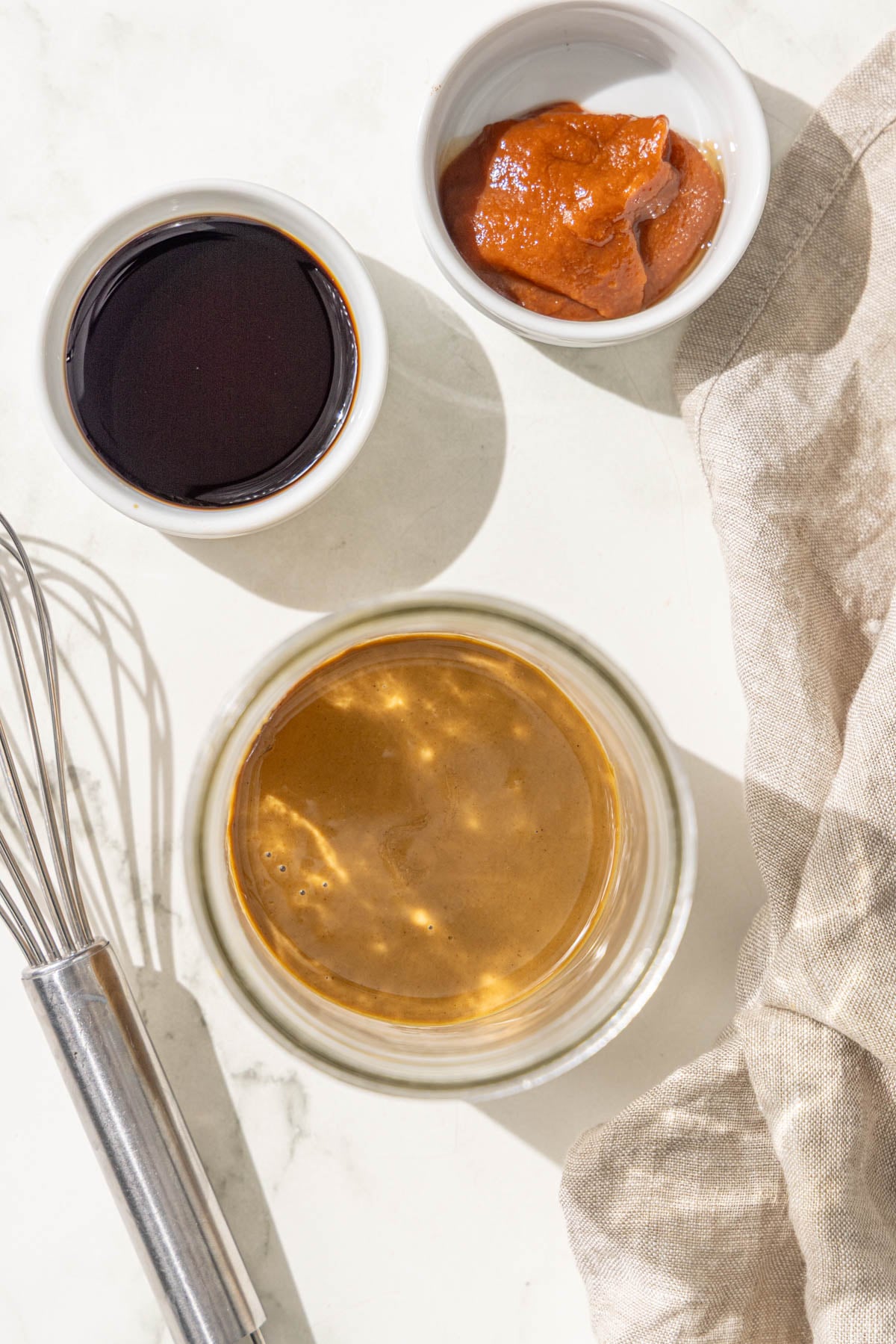 A jar of sesame paste, a bowl of soy sauce, a bowl of sriracha, and a mini whisk.