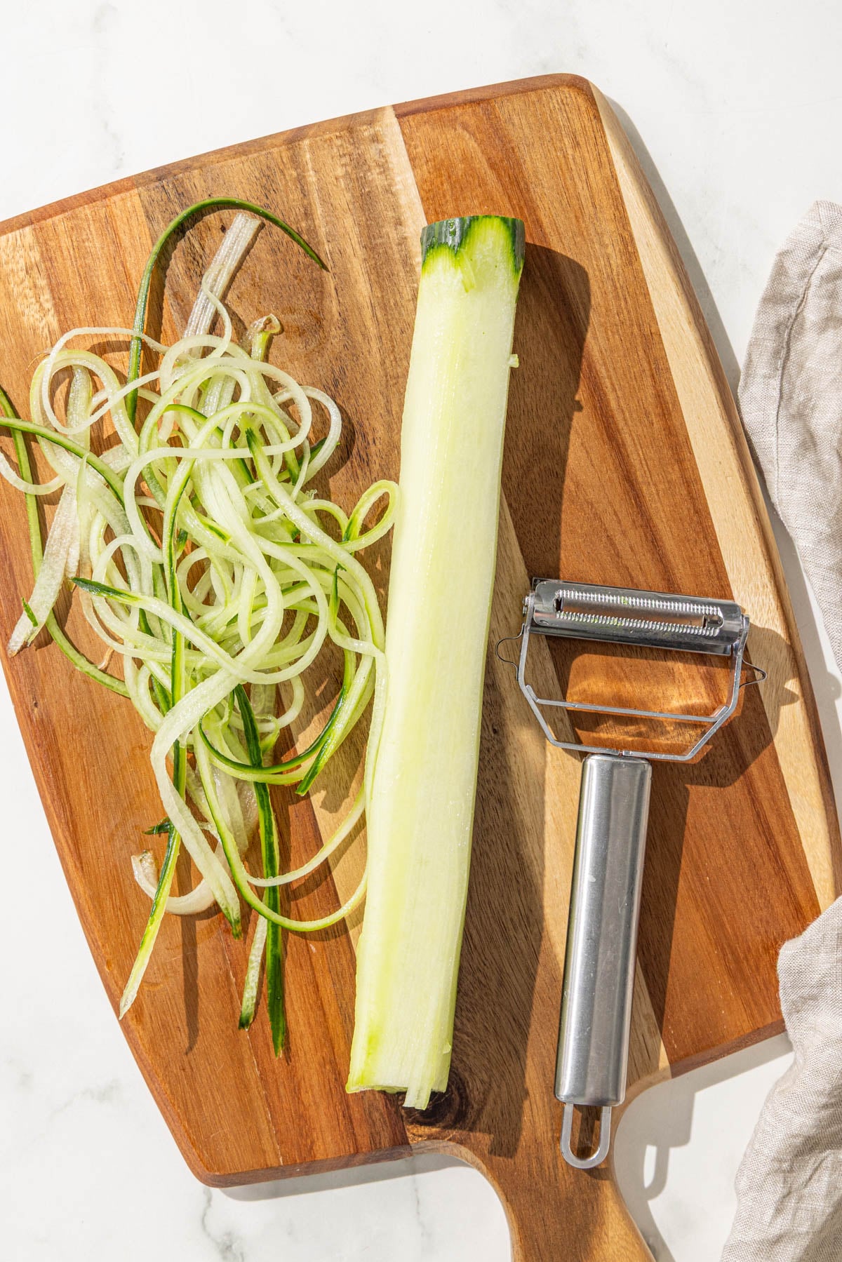 A wooden cutting board with julienned cucumber and a peeler on it.