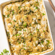 A 9x13 spinach cheesy quinoa casserole topped with parsley and breadcrumbs and a wooden spoon on the counter next to the dish.